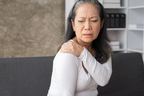 A woman grabs her shoulder with a look of discomfort on her face.