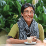 Professor Thomas sits in front of a leafy plant at a table smiling at camera with her hands clasped in front of her hot beverage