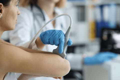 A woman in a white coat and blue latex gloves rolls a scanner along a woman's arm
