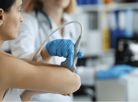 A woman in a white coat and blue latex gloves rolls a scanner along a woman's arm
