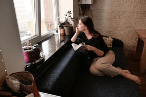 young woman sitting in a window seat with a book and looking out of the window, thinking