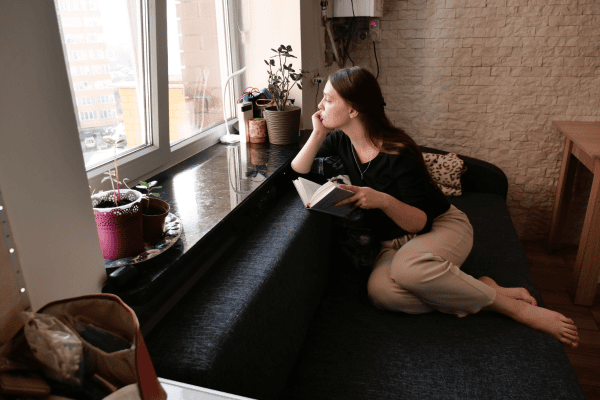 young woman sitting in a window seat with a book and looking out of the window, thinking